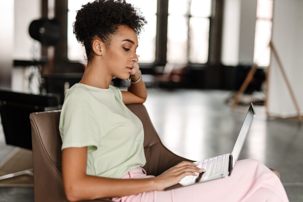 Young black woman working with laptop while sitting on armchair